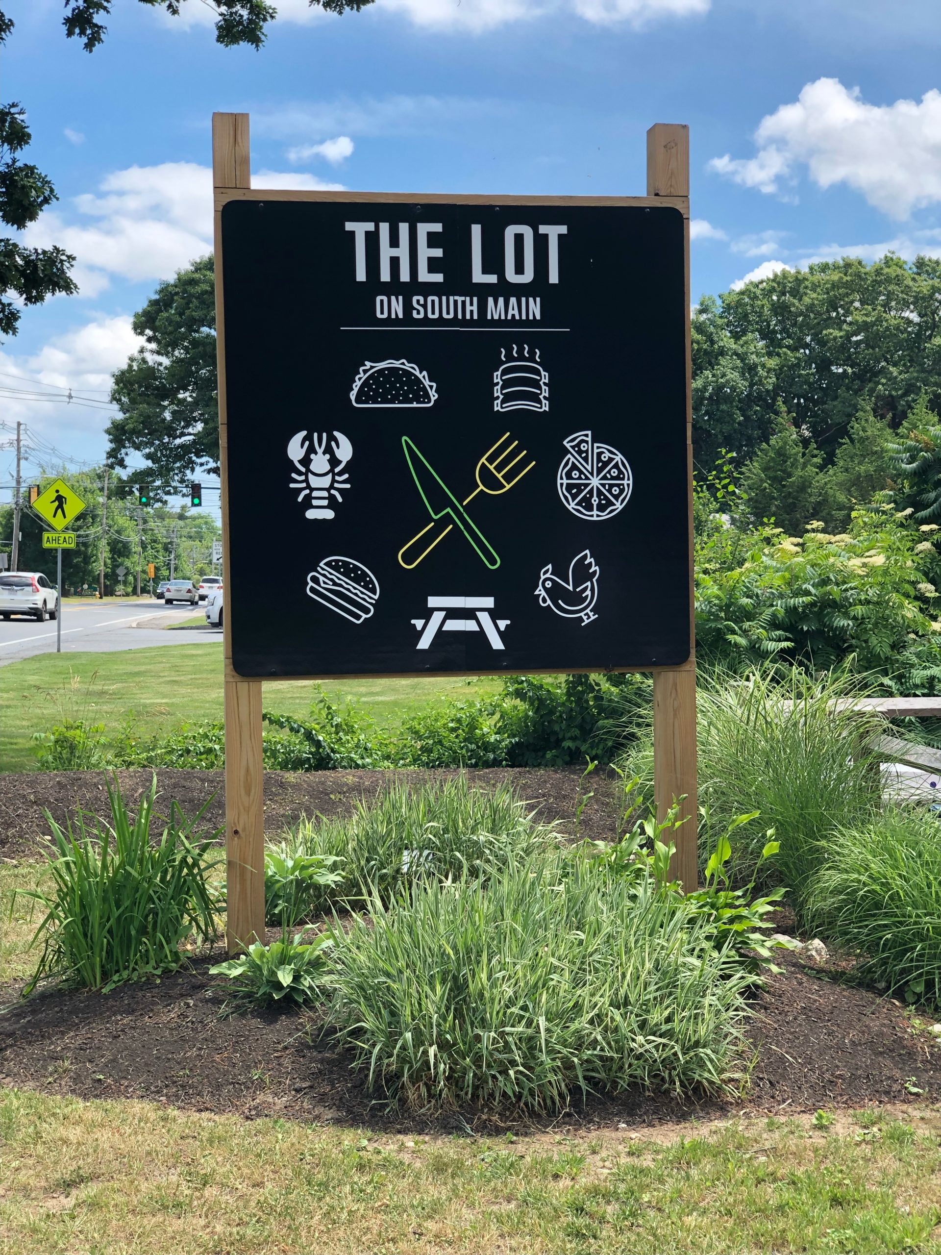 A sign titled "the lot on south main" with pictograms suggesting food, outdoor seating, and entertainment, backed by a green landscape and a clear sky with some clouds.