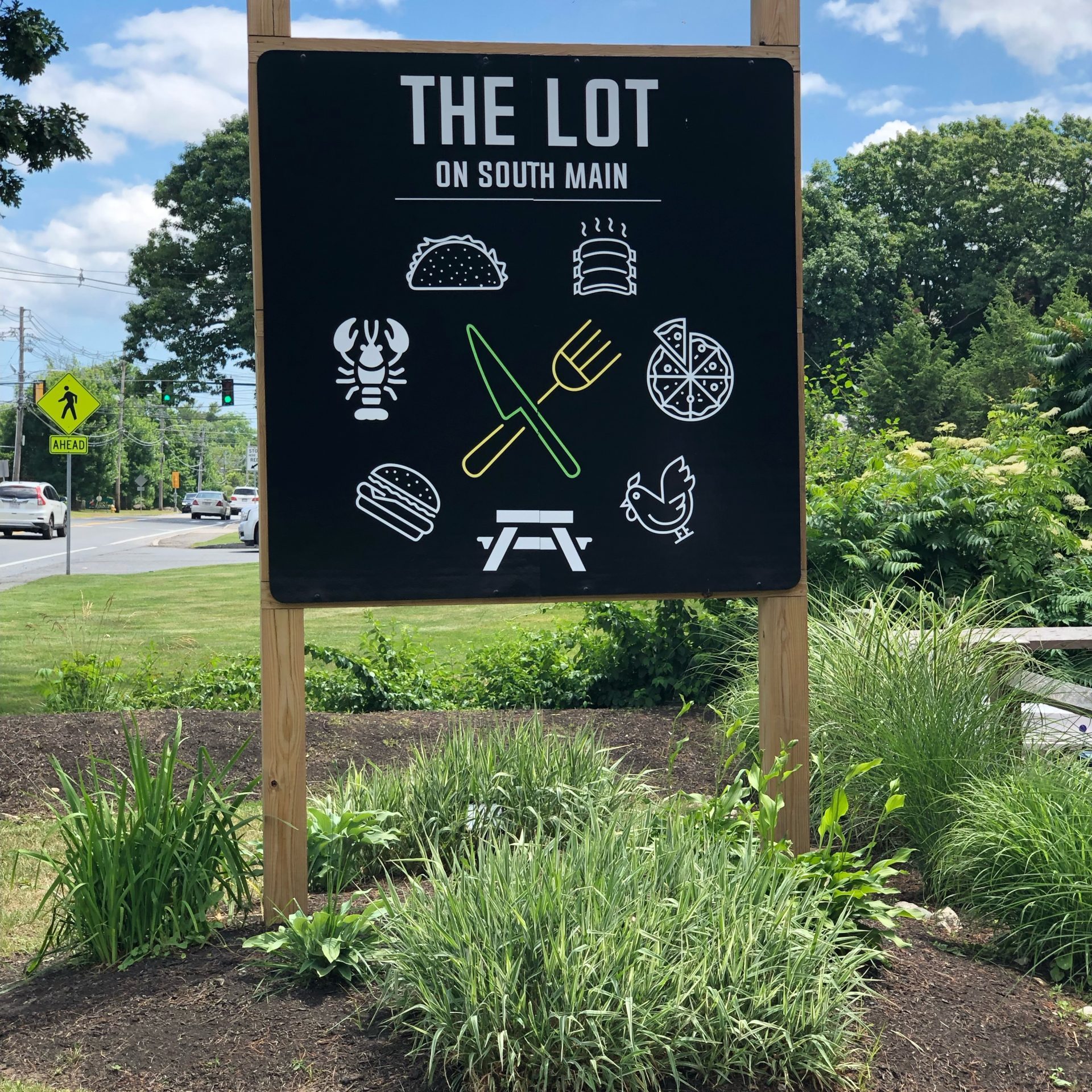 A sign titled "the lot on south main" with pictograms suggesting food, outdoor seating, and entertainment, backed by a green landscape and a clear sky with some clouds.